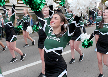 Michigan State pompon team marching in a parade, in their green and black uniforms and green and white pompons in the air.