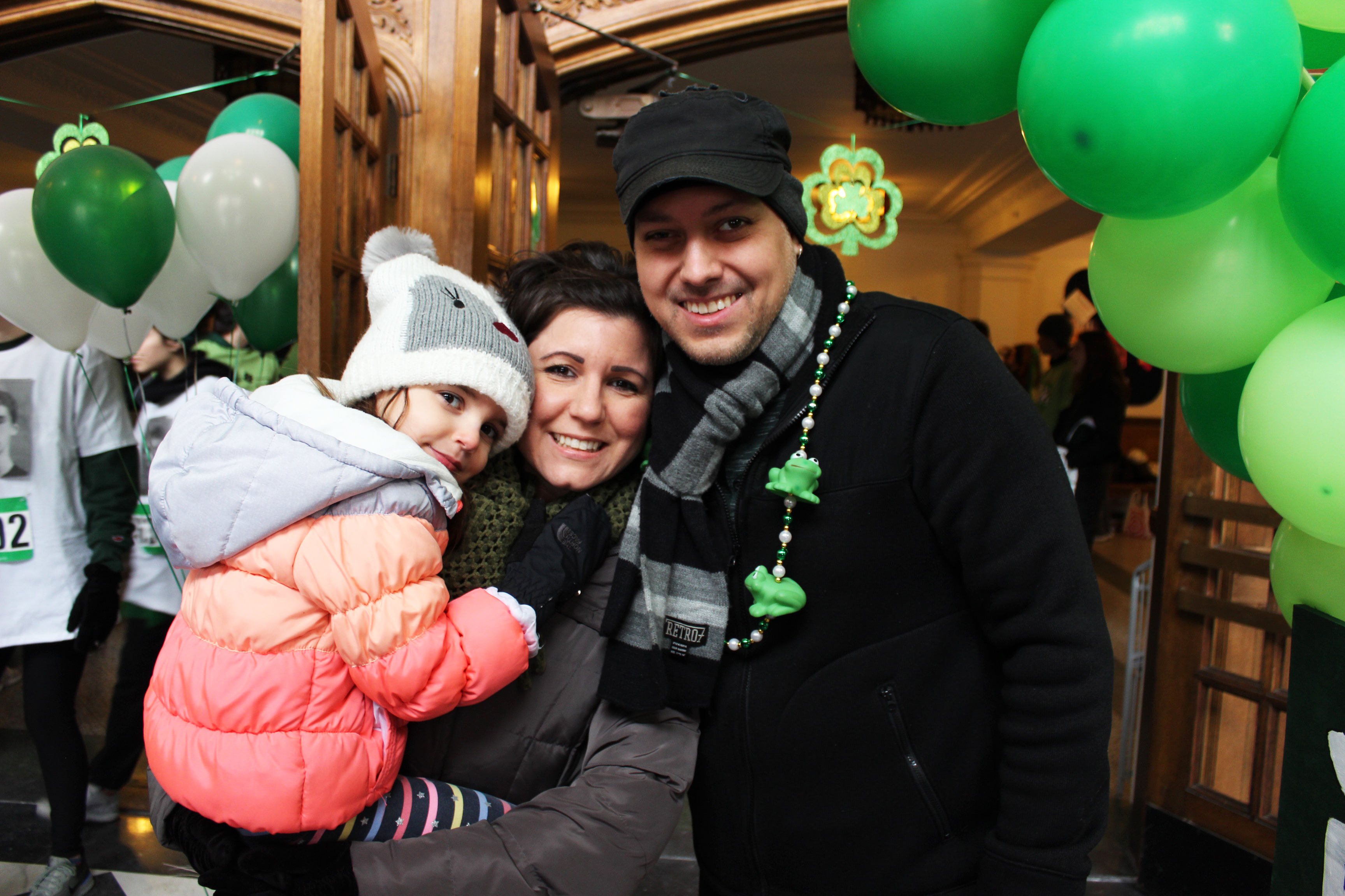 Photo of Caleb Sandoval with his wife Leanne and daughter Charlotte at the 19th Annual RCPD/Tower Guard Shamrock 5k Race in 2019 posing in front of Shamrock decorations and green and white balloons. 