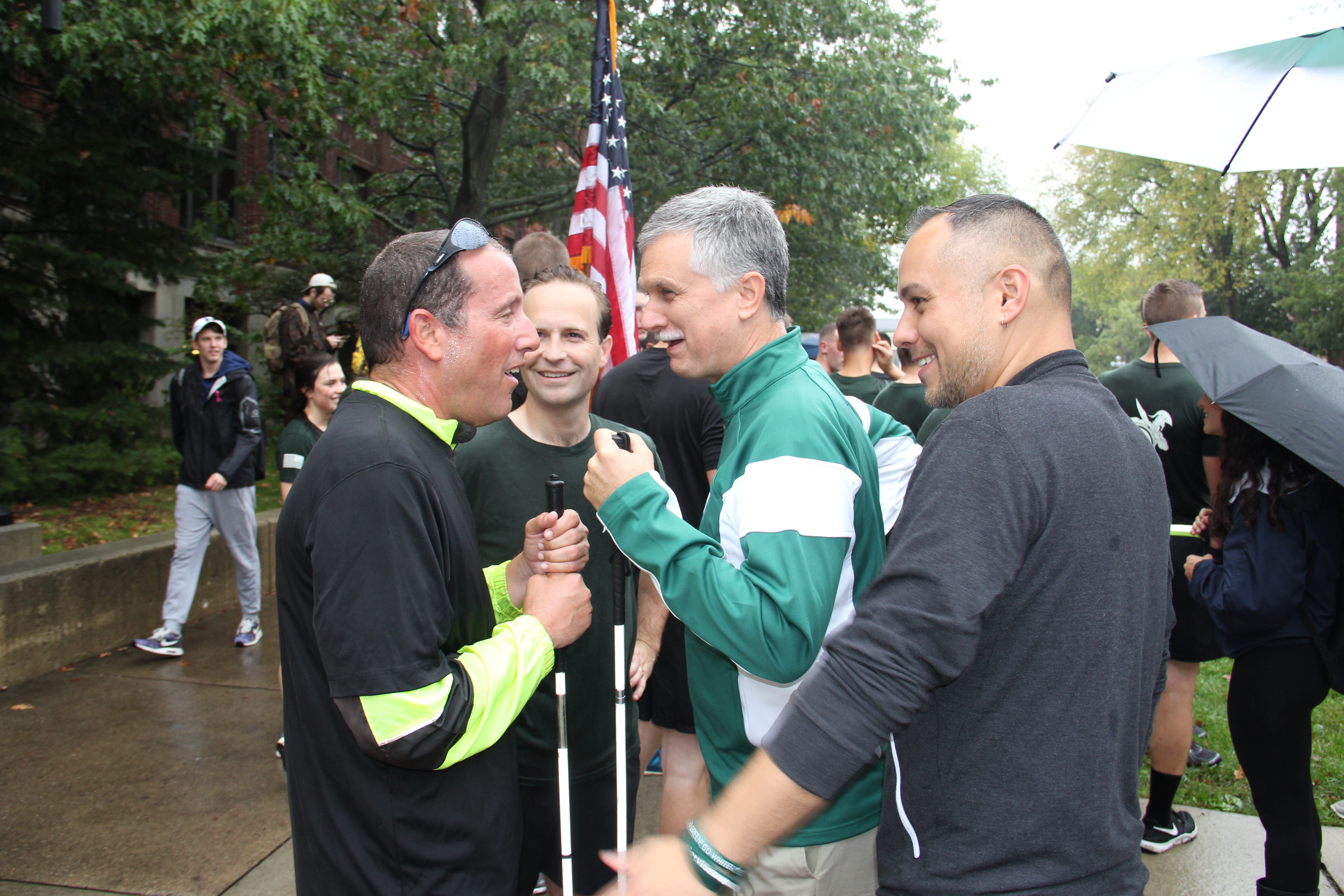 Candid photo of Justice Richard Bernstein, Fmr. Lt. Gov. Brian Calley, Michael Hudson and Caleb Sandoval at the Alex’s Great State Race in 2017, all four are standing in conversation and smiling, ROTC Cadets are visible in the background holding the American flag.