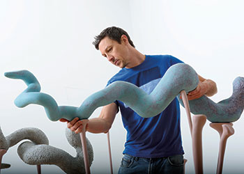 MFA student Joe Wilkinson carefully places a coiled clay sculpture, painted blue, atop flesh-toned wooden spikes as part of his art installation.