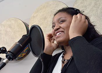 A young girl wearing a headset sings into a microphone in a recording studio. She is smiling, wearing a black sweater and a beaded necklace.