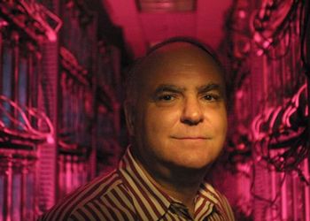 John R. Koza wears a striped shirt and stands in a pink-lighted hallway full of computer circuit boards.