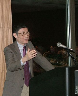 The late Professor Tung stands at a podium and speaks into a microphone.