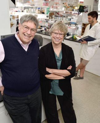 Dan and Karen Friderici pose in a lab with a student in a labcoat working in the background