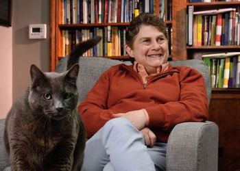 Sue Levy sits in a chair, with bookshelves in the background and her cat in the foreground