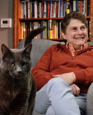 Sue Levy sits in a chair, with bookshelves in the background and her cat in the foreground