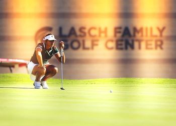 A golfer lines up her shot with the Lasch building in the background