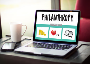 A computer with the word philanthropy and ways to donate on it sits next to a coffee cup and a cellphone on a desk
