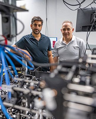 Elad Harel and Marcos Dantus pose behind cables and optical equipment in a lab space.