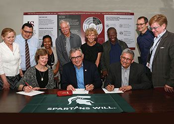 leaders from universities sign a paper at a table with a spartan flag