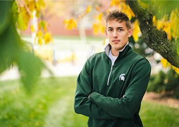 MSU student Jordan Weber faces the camera dressed in Spartan green, standing on a campus lawn with trees in the background.