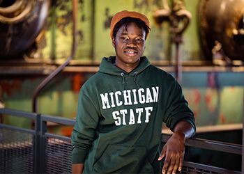 MSU student Kwaku Baffour-Awuah faces the camera dressed in Spartan green, standing against a gray and green backdrop inside the STEM Teaching and Learning Facility.