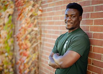 MSU student Kofi Debrah faces the camera dressed in Spartan green, standing outside against a red brick wall.