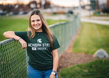 MSU student Jordan Weber faces the camera dressed in Spartan green, standing outside against an fence.