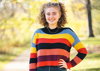 MSU student Maggie Lupton faces the camera dressed in colorful striped sweater, standing on a campus pathway with trees in the background.