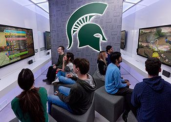 Students seated back-to-back at gaming consoles play a video game against each other.
