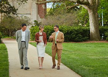 John Jiang, Karen Perry, and Rafael Auras, stroll along the sidewalk, chatting, with Beaumont Tower in the background.