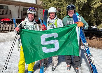 MSU alumni Jay Kirksey, Jay Horton, and Eric Reinhard hold a Spartan flag at the top of a ski hill.