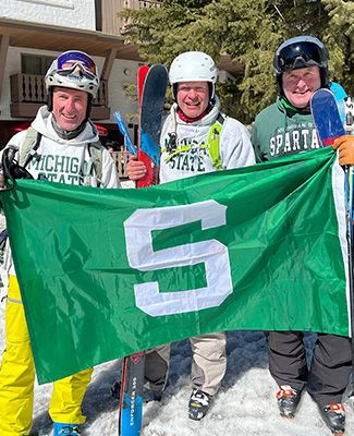 MSU alumni Jay Kirksey, Jay Horton, and Eric Reinhard hold a Spartan flag at the top of a ski hill.