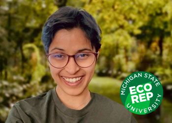 Veona Cutinho, a student with dark hair and wearing glasses and a green shirt, smiles in front of a forest.