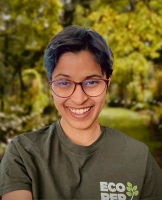 Veona Cutinho, a student with dark hair and wearing glasses and a green shirt, smiles in front of a forest.