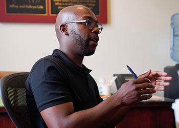 Photo of Howard Akumiah, a man with a short beard, wearing glasses and a black polo shirt, holding a pen in his hand as he talks to a person out-of-frame.