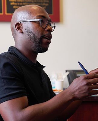 Photo of Howard Akumiah, a man with a short beard, wearing glasses and a black polo shirt, holding a pen in his hand as he talks to a person out-of-frame.