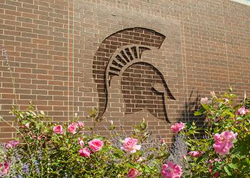 The MSU Spartan helmet logo set into the bricks on the facade of Breslin Center with flowers underneath.