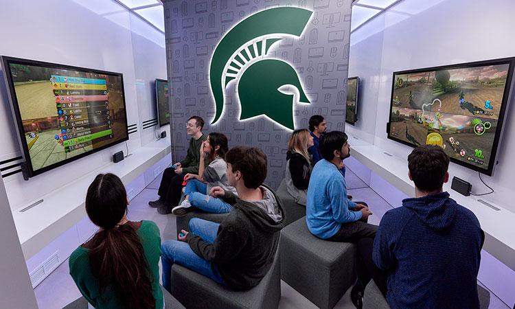 The Spartan helmet is visible in the background of a photo of students playing a multiplayer game on large TV screens.