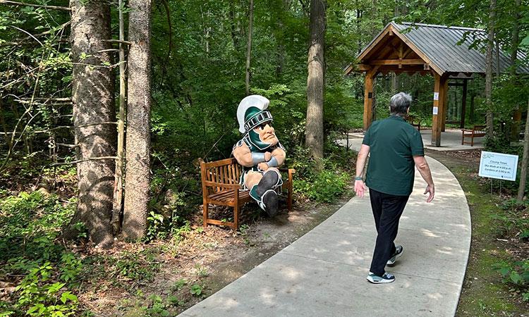 Sparty sits on a bench as a person walks down the Gruel Nature Trail