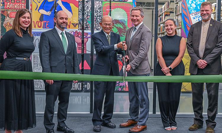 Two people are cutting a large ribbon with large scissors while others stand beside them. There are shelves of books in the background. Colorful murals are at the end of each book stack.