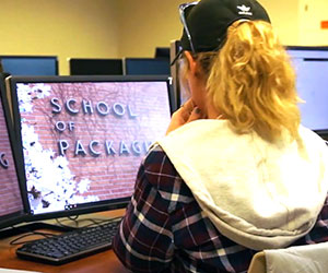 Student sitting at computer with screen showing "School of Packaging" letters on brick wall