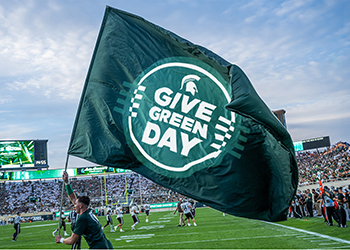 Spartan cheerleader running carrying a large green flag with Give Green Day letters in white