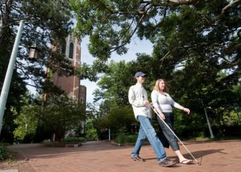 Two individuals walking on campus, one has a white cane for assistance with visual imparements 