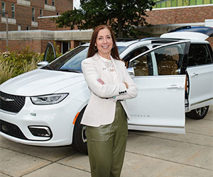 Chris Feuell, in green pants and a white suit coat, in front of an MSU building and the donated, white, Chrysler Pacifica.